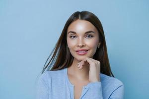 Confident pretty female entrepreneur cares about her complexion, keeps hand under chin, enjoys productive lucky day, listens information carefully, stands against blue background. Sweet cute look photo