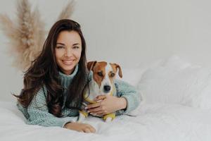 Photo of cheerful woman embraces dog with love, spend free time together, expresses tender feeling and emotions, falls in love with pet, lie on comfortable bed. Positive emotions, animals and care