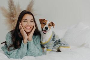 Horizontal shot of lovely brunette female in casual wear, lies on white bedclothes with pedigree dog, expresses happiness, enjoys company of pet, rest together at home, feel relaxed and satisfied photo