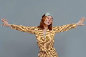 Positive relaxed woman wears blindfold and striped robe stretches arms sideways, smiles broadly, closes eyes, isolated over grey background, wakes up in bedroom, slept well. Good morning concept photo