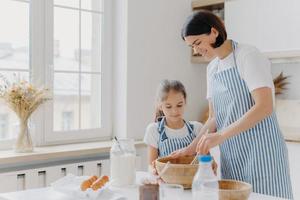 Photo of happy mum and child cook together at kitchen, wears aprons prepare something tasty, make food, whisk eggs with beater, bake house pastries at home. Milk, eggs, chocolate, flour on table