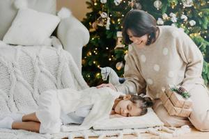 Attractive brunette smiling woman stands on knees with present boxes, little daughter sleeps near on pillow, has pleasant dreams, enjoy holiday. Winter time, New Year and Christmas. Rest concept photo