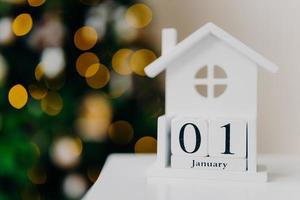 vacaciones de invierno, decoración y primer día del año. casa de madera blanca creativa hecha a mano con fecha escrita que simboliza año nuevo, luces de guirnalda foto