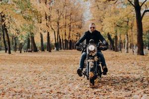 Brutal professional male motorcyclist rides bike, wears sunglasses, gloves and black jacket, has ride through autumnal park, beautiful scenery in background with yellow trees and fallen leaves around photo