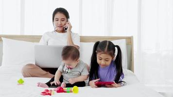 el bebé y la pequeña hija juegan al juguete en la cama mientras la madre independiente ocupada trabaja en la computadora portátil