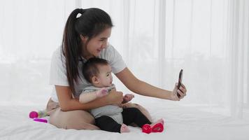 Aziatische moeder kijkt naar de smartphone met haar baby op bed video