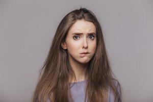 Offended stressful female bites lower lip with dissatisfaction, has sorrorful expression, expresses negative emotion, being in low spirit after quarrel with somebody, isolated over grey background photo