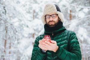 Attractive serious male wears warm winter clothes, keeps candle, stands against winter forest background, enjoys fresh air, being very confident. Fashionable unshaven man outdoors. photo