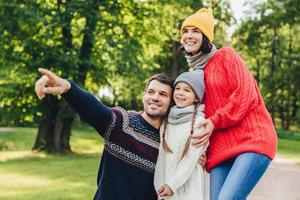 tres miembros de la familia pasan tiempo juntos, miran el hermoso lago en el parque, indican con los dedos, están de buen humor, sonríen agradablemente. padre, madre e hija disfrutan de la unión, ambiente tranquilo foto