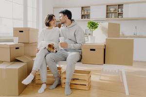 Lovely spouses sit near carton big boxes, drink coffee and talk about future plans, relocate in new apartment, pose with pedigree dog, poses in spacious kitchen, think about design or interior photo