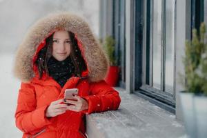 Smiling young European female in red coat, has black manicure, uses modern electronic gadget for communication in internet with friends, likes winter weather, sends photo in social networks.