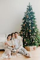 Three family members pose on floor in cozy room, gift boxes around, decorated New Year tree and sofa. Father, mother and daughter with jack russell terrier dog anticipate for coming holiday. photo