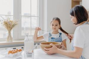 Shot of small kid helper whisks cream with beater, learns how to bake, listens useful advice from mother who holds her pony tail, sit at white kitchen table together. Family cooking together photo
