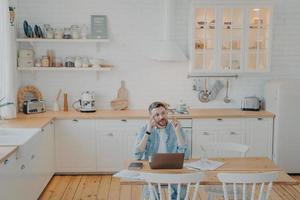 Young office worker working remotely from home with computer while sitting by kitchen table photo