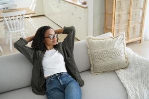 Tranquila joven afroamericana sentada relajándose soñando en un acogedor sofá en la sala de estar en casa foto