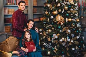 Beautiful brunette female embraces her little daughter and her husband sit together near decorated New Year tree, being glad to spend holidays in family circle. Affectionate family with happy looks photo