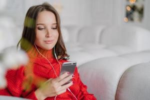 Beautiful young woman holds cell phone, listens music from playlist, surfes social networks and messages with friends, enjoys calm atmosphere at home, sits on comfortable sofa. Lifestyle concept photo