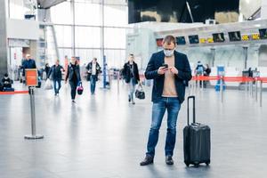 Horizontal shot of man poses in crowded airport, uses cell phone, checks time of flight online, stands near suitcase, wears medical mask during coronavirus crisis. Health care, traveling concept photo
