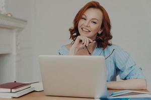 Self employed female freelancer has gentle smile on face, works on startup project, poses with laptop computer at desktop, searches information for productive work, sits indoors, holds eyewear photo