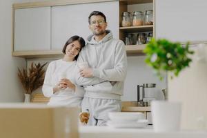 Indoor shot of pleasant looking wife leans at shoulder of husband, hold takeaway coffee, pose in kitchen, jack russell terrier dog poses near, cardboard boxes with personal stuff, modern interior photo