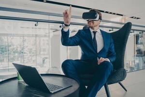 Well-groomed CEO man in formal suit wearing VR headset goggles, while sitting in office lobby photo