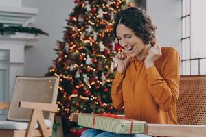 Excited Italian woman sitting on sofa near Christmas tree at home with wrapped xmas gift box photo