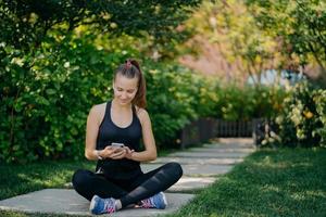 Outdoor shot of athletic woman rests after workout sits crossed legs in park surfs social networks dressed in sportswear listens music via wireless earphones uses sport app enjoys good weather photo