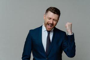 Positive businessman clenches fist exclaims from joy hooray celebrates success dressed formally satisfied after successful business deal isolated over grey background. Congratulations I did it photo