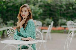 Sideways shot of beautiful Caucasian woman in elegant clothes, wears makeup, holds mobile phone, spends free time in outdoor cafe, waits for friend. People, lifestyle, rest and technology concept photo