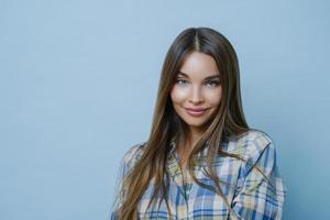 Portrait of good looking female student poses against blue studio wall, looks at camera with charming smile, dresses checkered shirt, poses for university photo, feels optimistic, wears make up photo