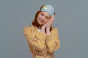 Pleased cheerful woman wears blindfold and striped pajamas, has happy expression, awakes in good mood, has healthy sleep habits, feels totally relaxed, smiles broadly, isolated on grey background photo