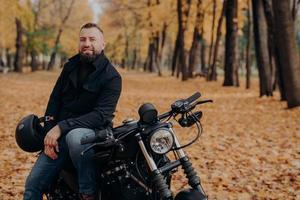 Bearded male motorcyclist rides black bike, holds helmet, has travel on his own transport, poses in park during fall season, looks happily at camera. Carefree biker enjoys journey or trip on vehicle photo