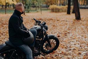 Back view of bearded male biker looks thoughtfully somewhere into distance, poses on black motorbike, holds protective helmet, spends leisure time in autumnal park, copy space for your advertising photo