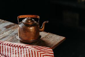 Horizontal shot of antique kettle on vintage wooden table. Old crockery. Metal copper teapot against dark background red towel near. Selective focus photo