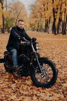 Brutal male biker rides motorcycle during autumn time, poses in park with many trees and yellow foliage, enjoys driving in open air, has tour journey. Motorcyclist with own transport outdoor photo