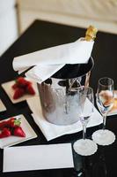 Bottle of champagne in bucket with ice and empty glasses on black table with delicious strawberry. Served table with dessert and alcoholic drink for special occasion photo