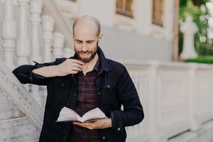 Handsome clever bald male holds book, reads interesting novel with serious expression, stands outdoor against ancient white building, likes reading. People, hobby, lifestyle and leisure concept photo