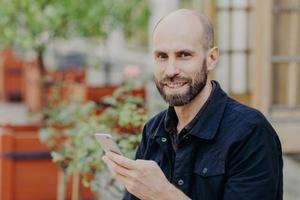 toma al aire libre de un hombre apuesto con barba oscura y bigote, de buen humor, pasa tiempo libre afuera, sostiene un teléfono inteligente, busca redes sociales, conectado a internet inalámbrico. foto