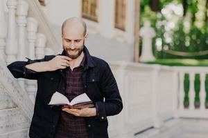 Clever middle aged student learns and cramms information before final exam, focused in book, has gentle smile, thick beard, poses outdoor near old building. People, leisure and education concept photo