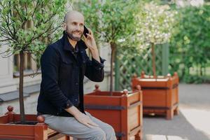 Attractive bald male with beard, has pleasant talk with girlfriend via cell phone, spends free time outdoor in park, being in good mood. Positive smiling young businessman has rest during weekends photo