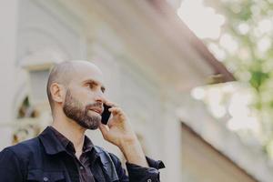 View from above of attractive middle aged male with beard, speaks with business partner via smart phone, poses against blurred background, enjoys using modern technologies. Man has conversation photo