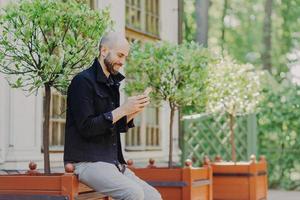 Concentrated male freelancer enjoys day off, breathes fresh air outdoor, uses modern cell phone, view photos in social networks has positive smile on face. Cheerful guy types text message on cellular