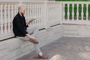 retrato al aire libre de un hombre feliz de mediana edad con una apariencia atractiva, sostiene un libro, lee algo divertido, disfruta del tiempo libre en el parque, posa contra el interior del balcón blanco. concepto de personas y pasatiempos foto