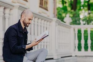 Sideways shot of bearded male with bald, holds book, sits outdoor against white balcony background, reads interesting detective story or scientific literature. People, education and hobby concept photo