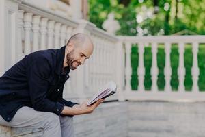 People, hobby and lifestyle concept. Postive bald man reads book with enthusiasm, poses outdoor against white balcony, has happy expression, enjoys calm atmosphere. Middle aged handsome male studies photo