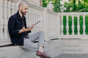 Attractive bald man reads attentively interesting book, gestures with hand, has stunning plot, tries understand feeling of main characters, poses outdoor, enjoys fresh air and wonderful view photo