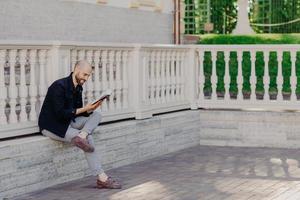 People, positive emotions and hobby concept. Unshaven bald middle aged man with happy expression, focused into book, reads something interesting, sits crossed legs, outdoor, has spare time. photo