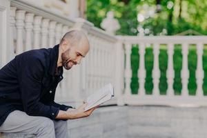 Photo of middle aged bald bearded man leans closely to book, has positive expression, sits against white balcony, has dark stubble, being in good mood as likes reading. Male reads romantic story