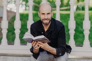 Positive bearded man looks directly into camera, reads very exciting and interesting book, sits against balcony background, spends spare time in summer garden. People, education and happiness photo