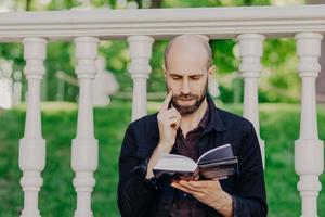 personas, estilo de vida, educación, concepto de hobby. atractivo hombre de mediana edad con rastrojo oscuro, sostiene un libro, se enfoca en la página, lee información informativa, posa al aire libre, tiene una expresión seria. foto